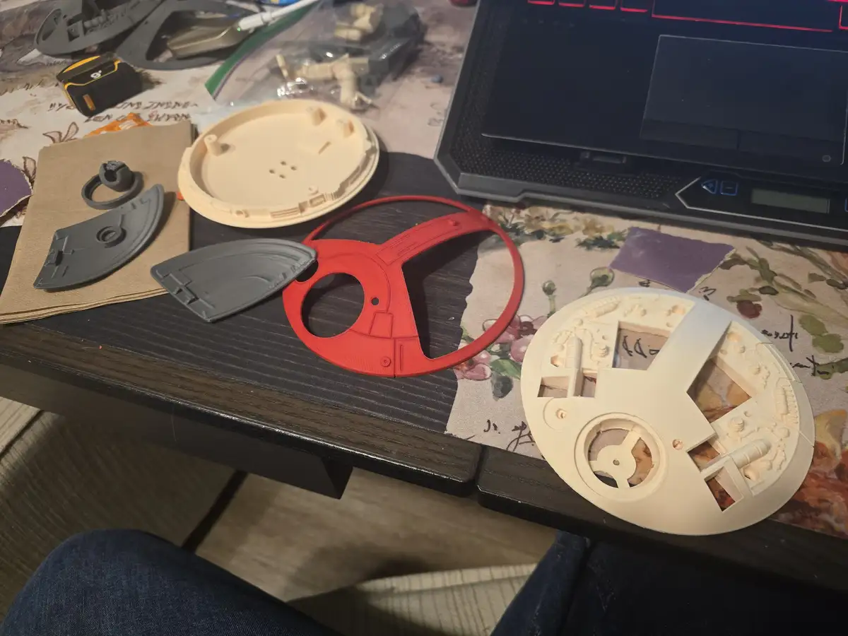 A cluttered desk shows various unassembled 3D-printed plastic parts, including a red ring-shaped piece and beige circular…