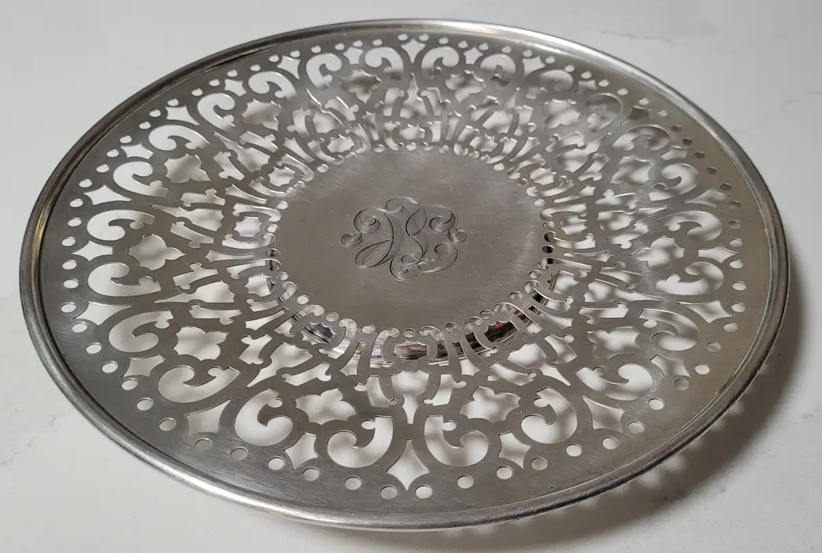 a close-up photo of a round, ornate silver tray with elaborate cut-out scrollwork patterns around its perimeter and a…