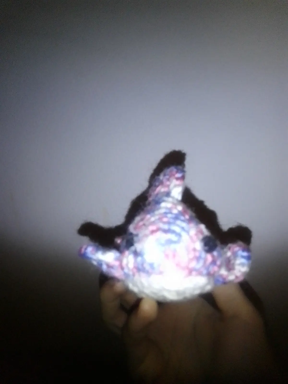 A blurry, low-light photo shows a hand holding a small, crocheted or knitted plush toy with a colorful, abstract pattern of…