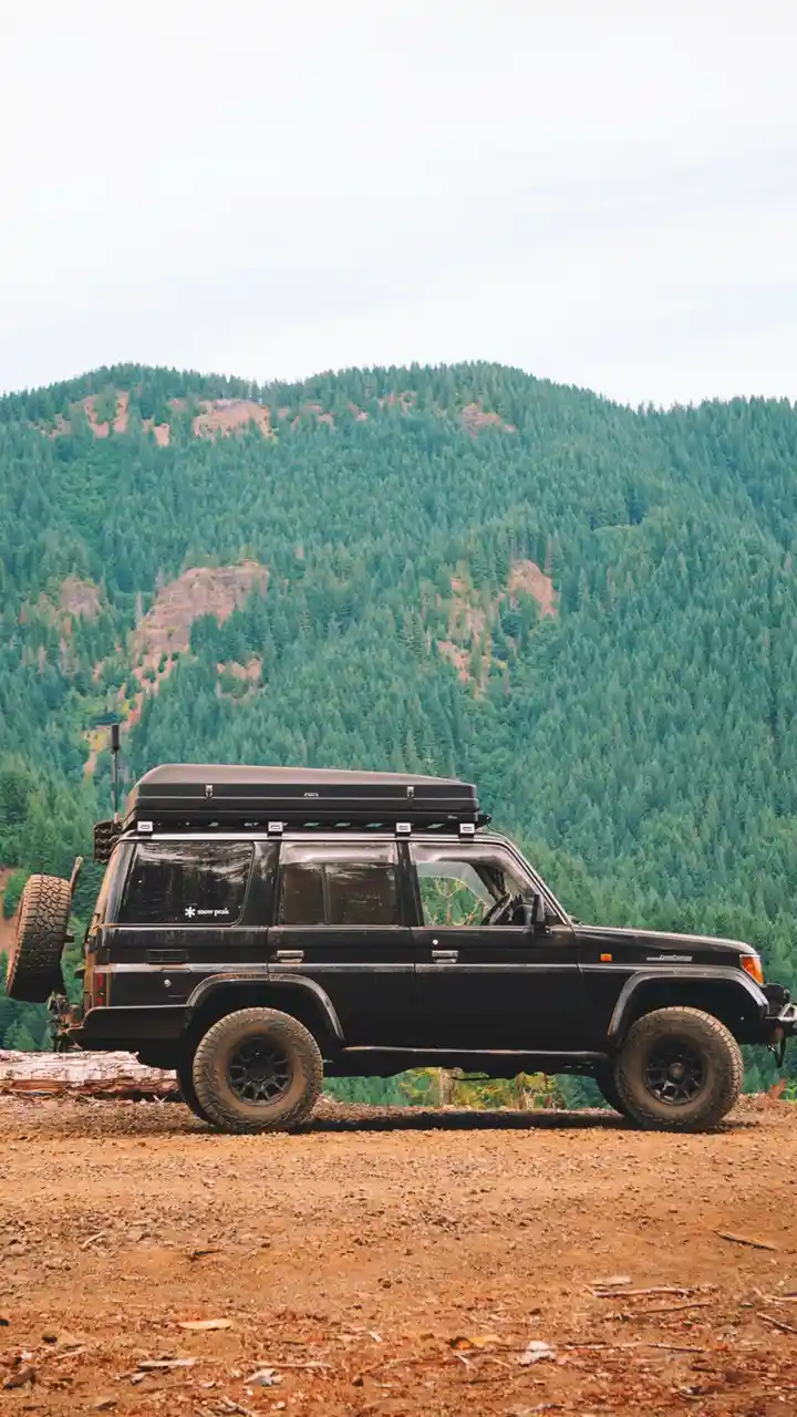 A black, rugged, older model SUV with a roof rack and spare tire mounted on the back is parked on a dirt road in front of a…