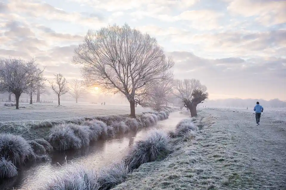 A person in a blue hoodie and dark pants runs along a frost-covered path beside a small stream in a winter park. Bare trees…