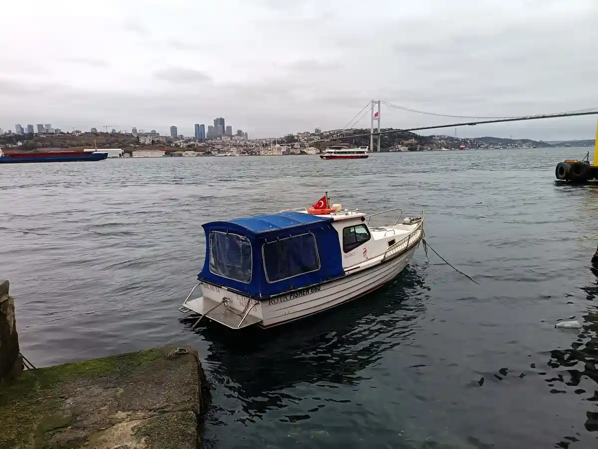 A small white and blue fishing boat named 'ROTA FISHER 660' is moored near a stone pier in a harbor, with a large suspension…