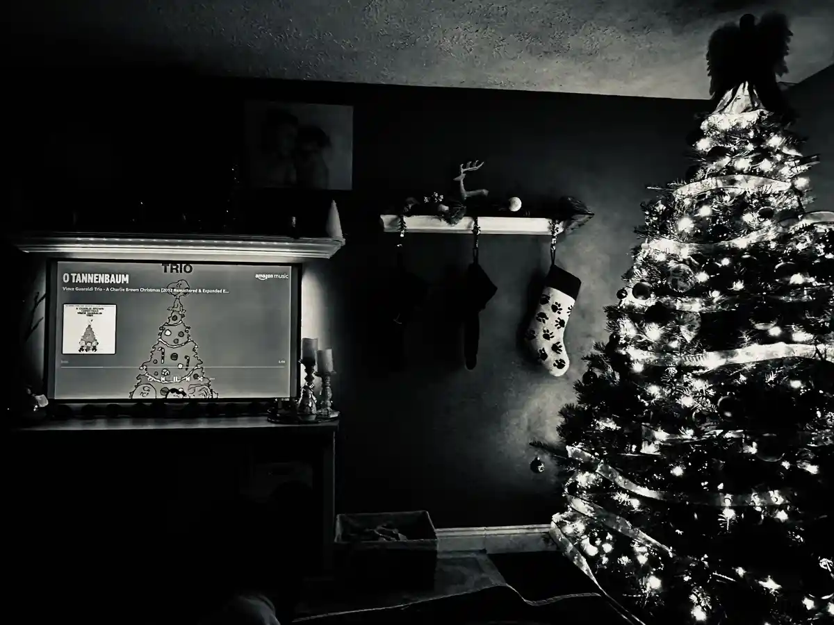 A black-and-white photo of a decorated Christmas tree next to a fireplace where a TV screen displays the song 'O Tannenbaum'…