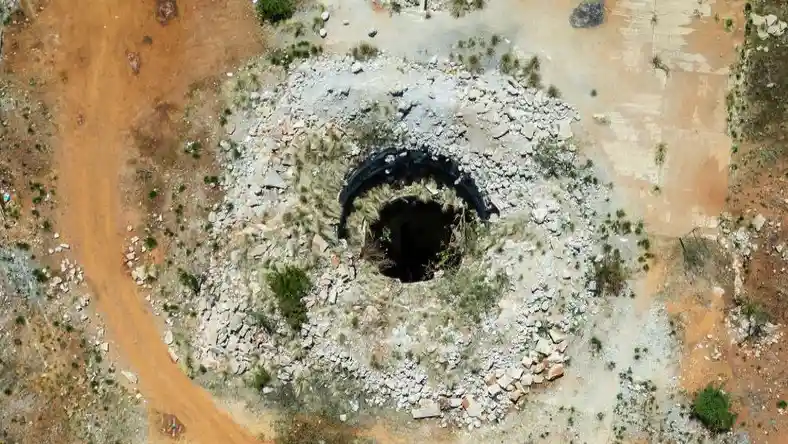 An aerial view shows a large, dark circular hole in the ground surrounded by scattered rubble, broken concrete, and sparse…