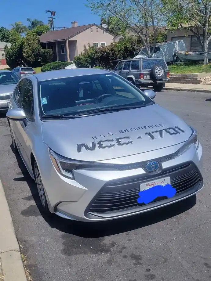 A silver Toyota Corolla is parked on a residential street with 'U.S.S. ENTERPRISE NCC-1701' written in black lettering…