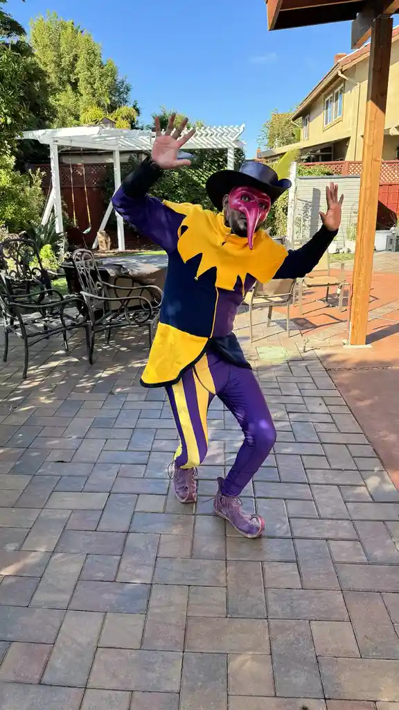 A person in a full jester costume with a long-nosed pink mask, purple and yellow outfit, and a black hat with a yellow…