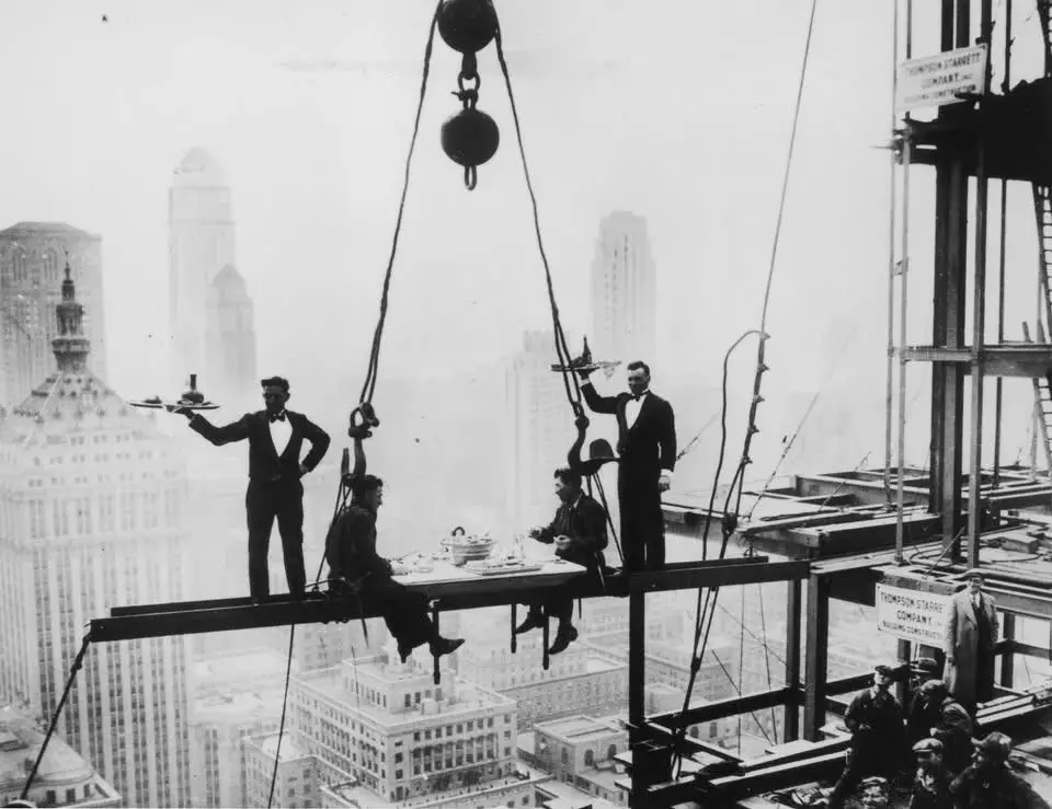 A vintage black-and-white photo shows workers on a high-rise construction site, with two men in tuxedos serving food and…