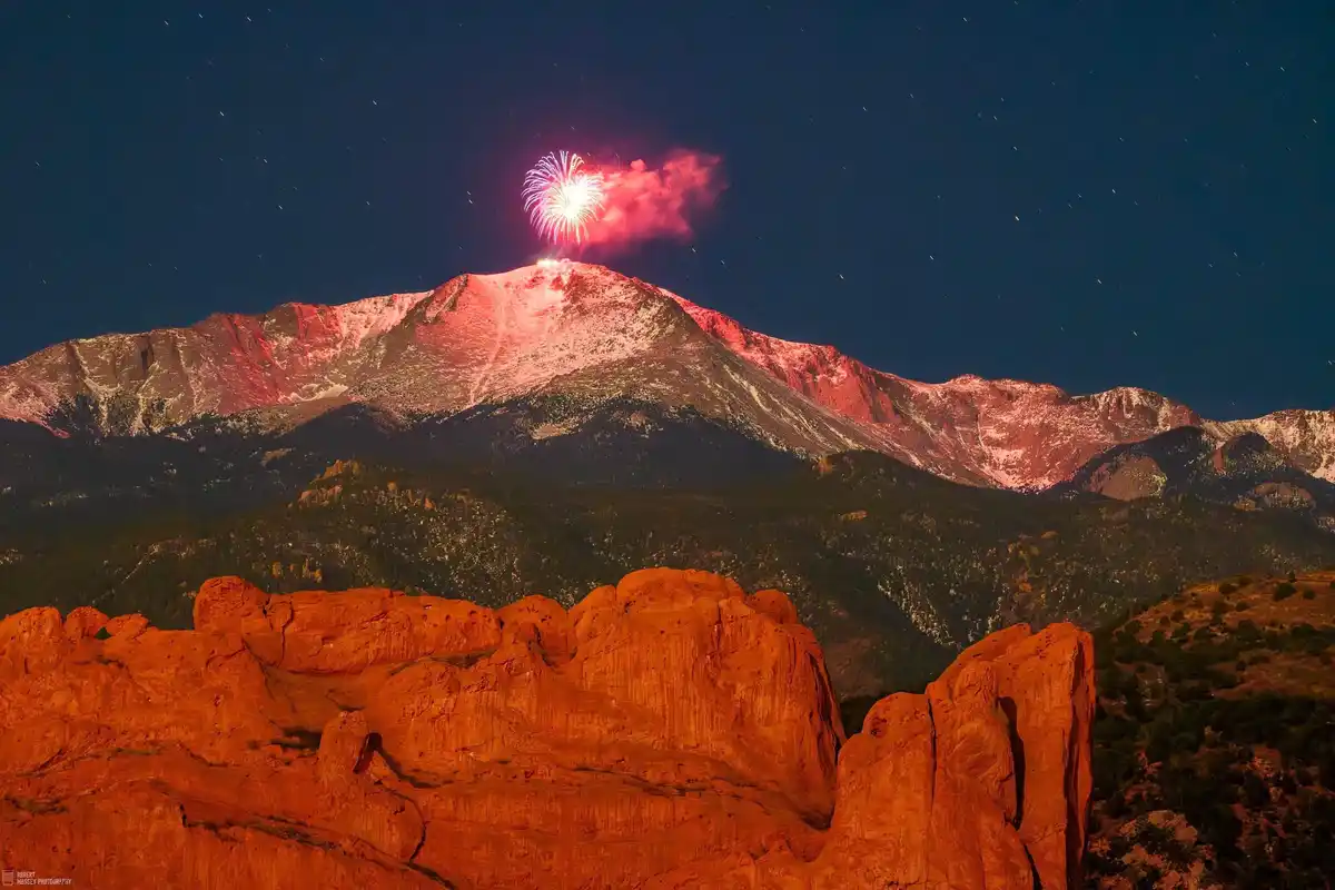 A nighttime photograph shows a majestic mountain range under a starry sky, with a bright fireworks explosion visibly…