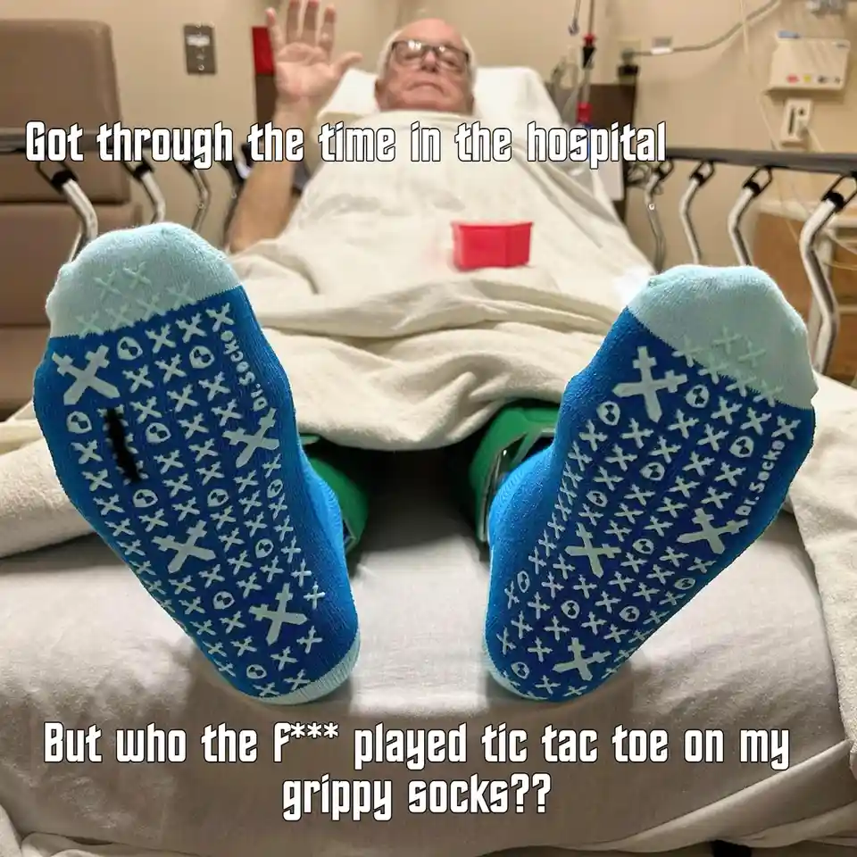 An elderly man lies in a hospital bed, raising his hand, with his feet in the foreground wearing blue socks patterned with…