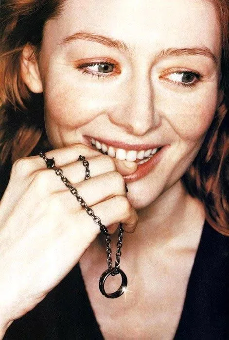 a woman with freckles and reddish hair smiles while holding a black chain necklace with a circular pendant near her mouth…