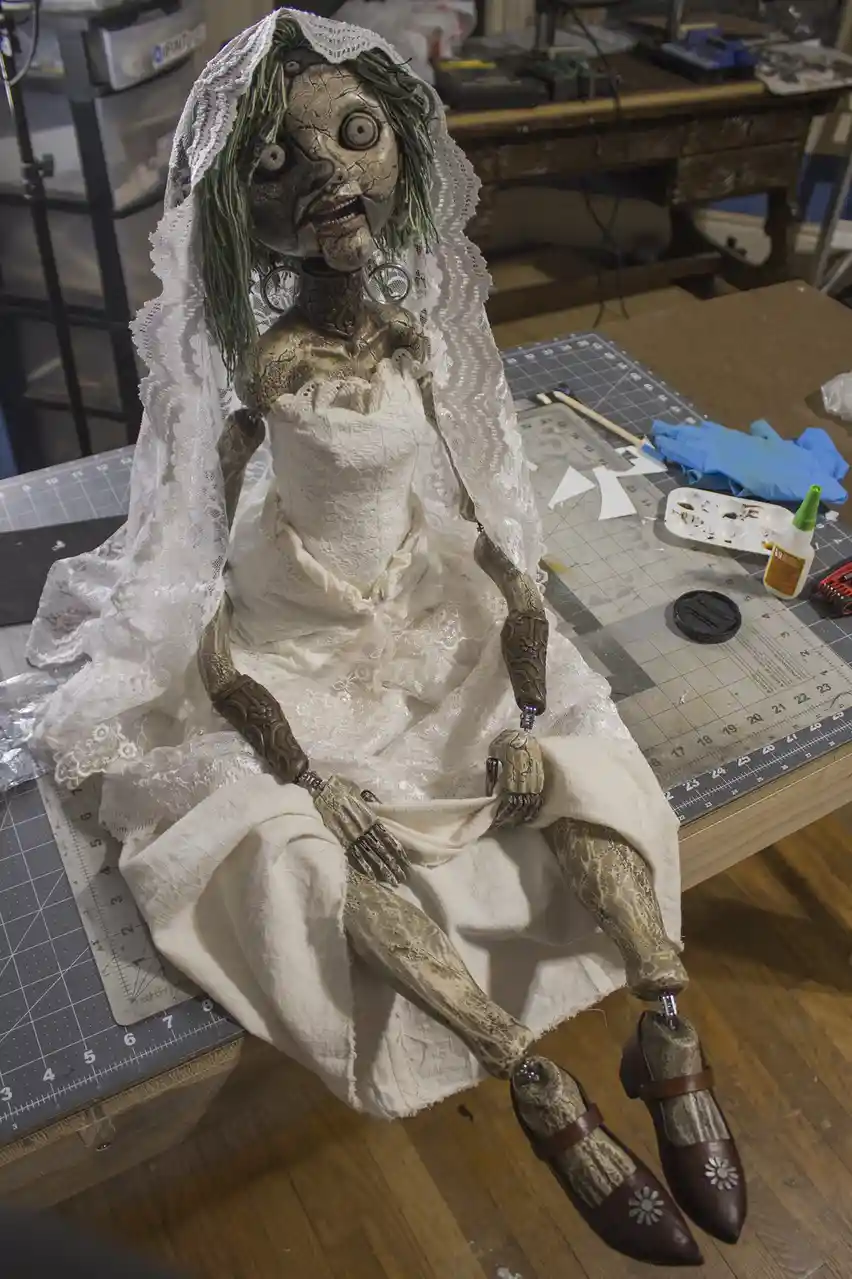 A grotesque, cracked-faced doll with green hair and a white lace veil sits on a workbench, wearing a tattered white wedding…