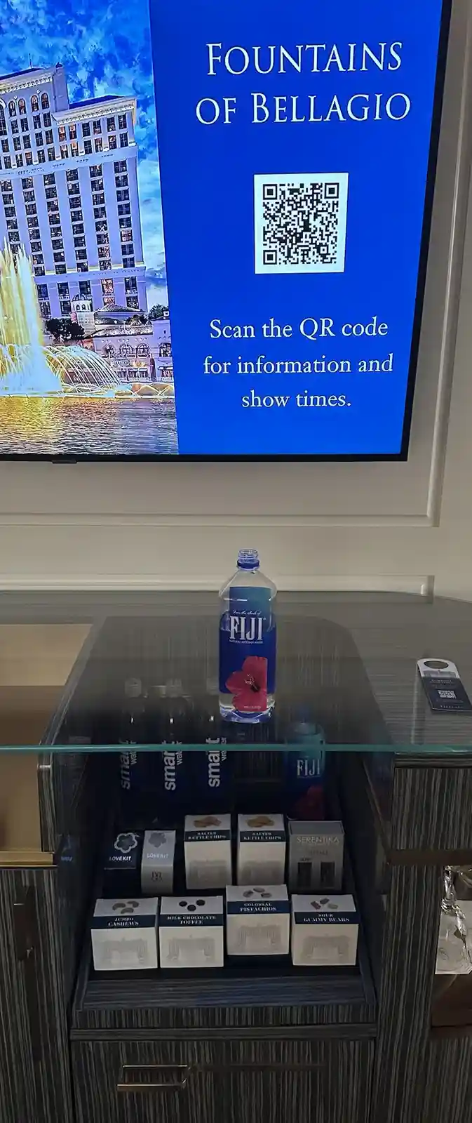A hotel minibar with Fiji water and snack boxes sits below a TV screen displaying 'Fountains of Bellagio' with a QR code and…