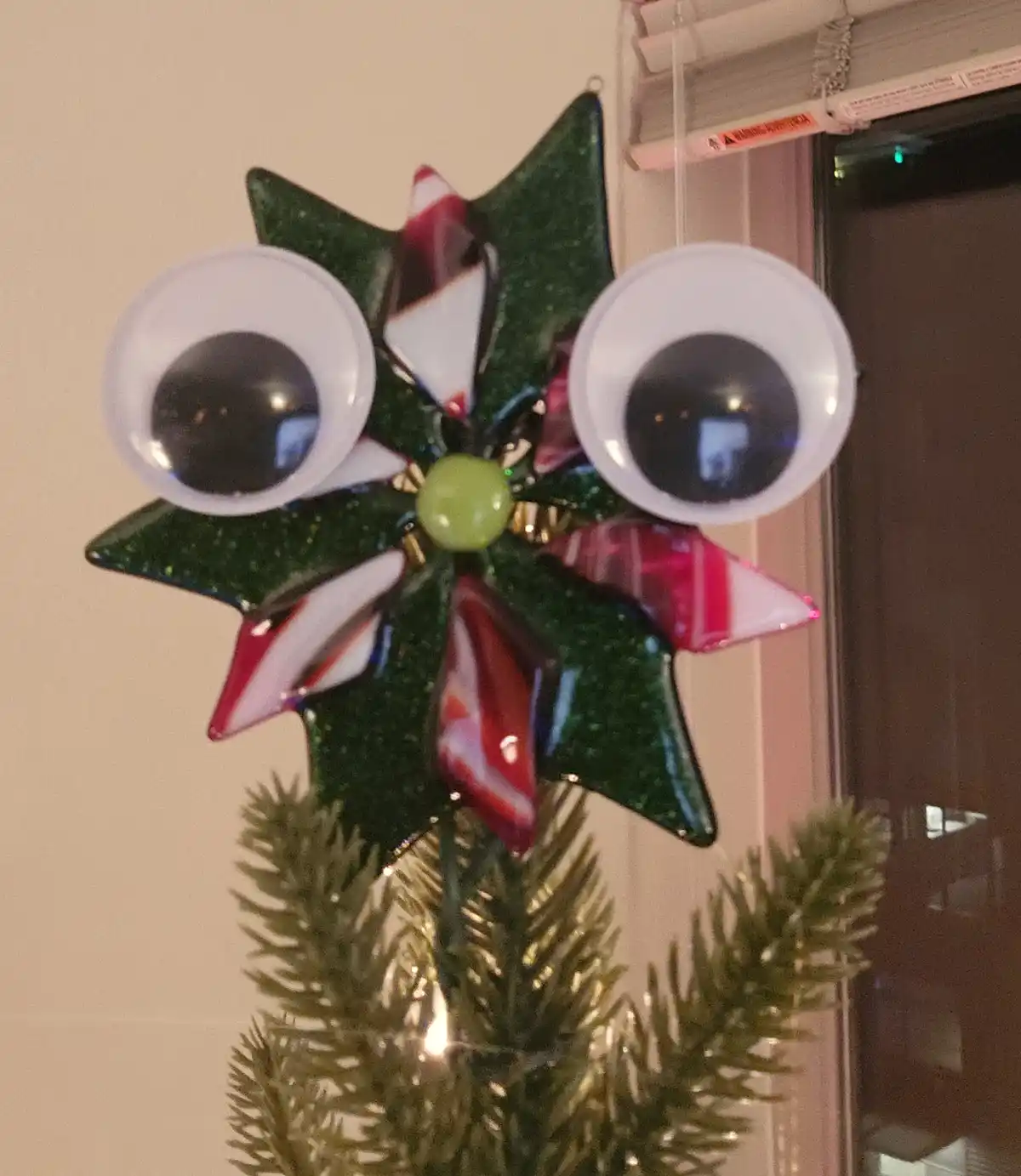 A handmade, glittery green star-shaped Christmas ornament with large googly eyes and red-and-white striped candy cane-like…