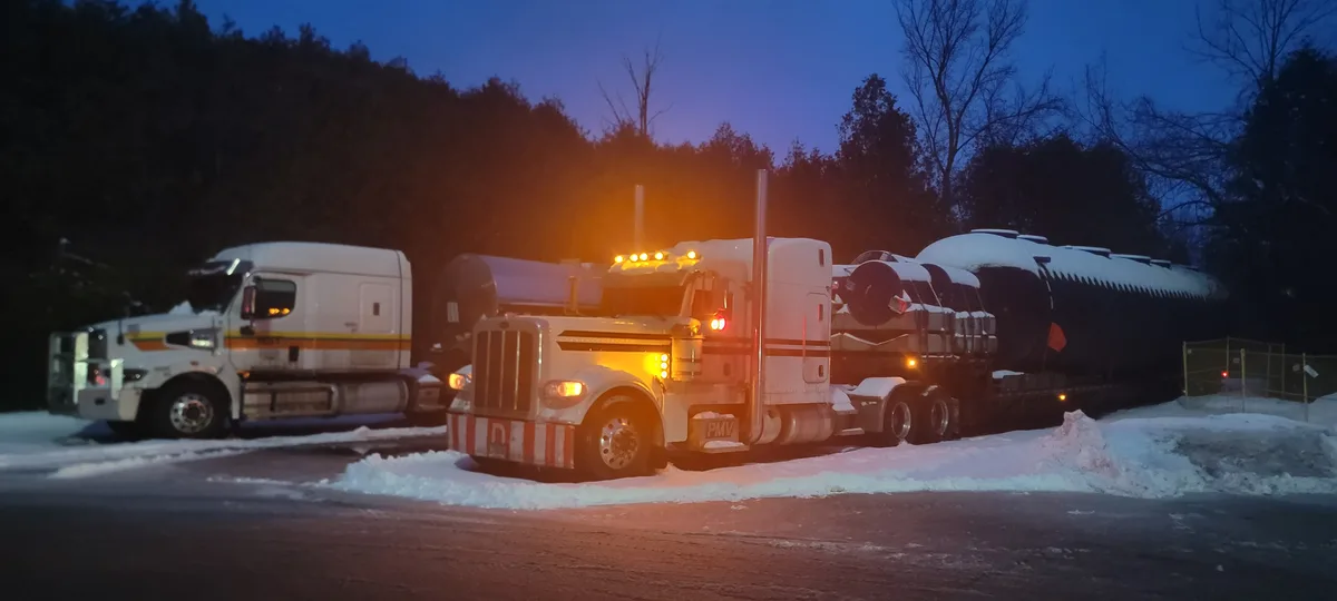 Two large white semi-trucks are parked side-by-side in a snowy lot at dusk, their headlights and warning lights glowing…