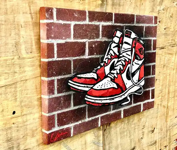 A canvas painting depicts a pair of red and white high-top sneakers, resembling Air Jordan 1s, resting against a brick wall…