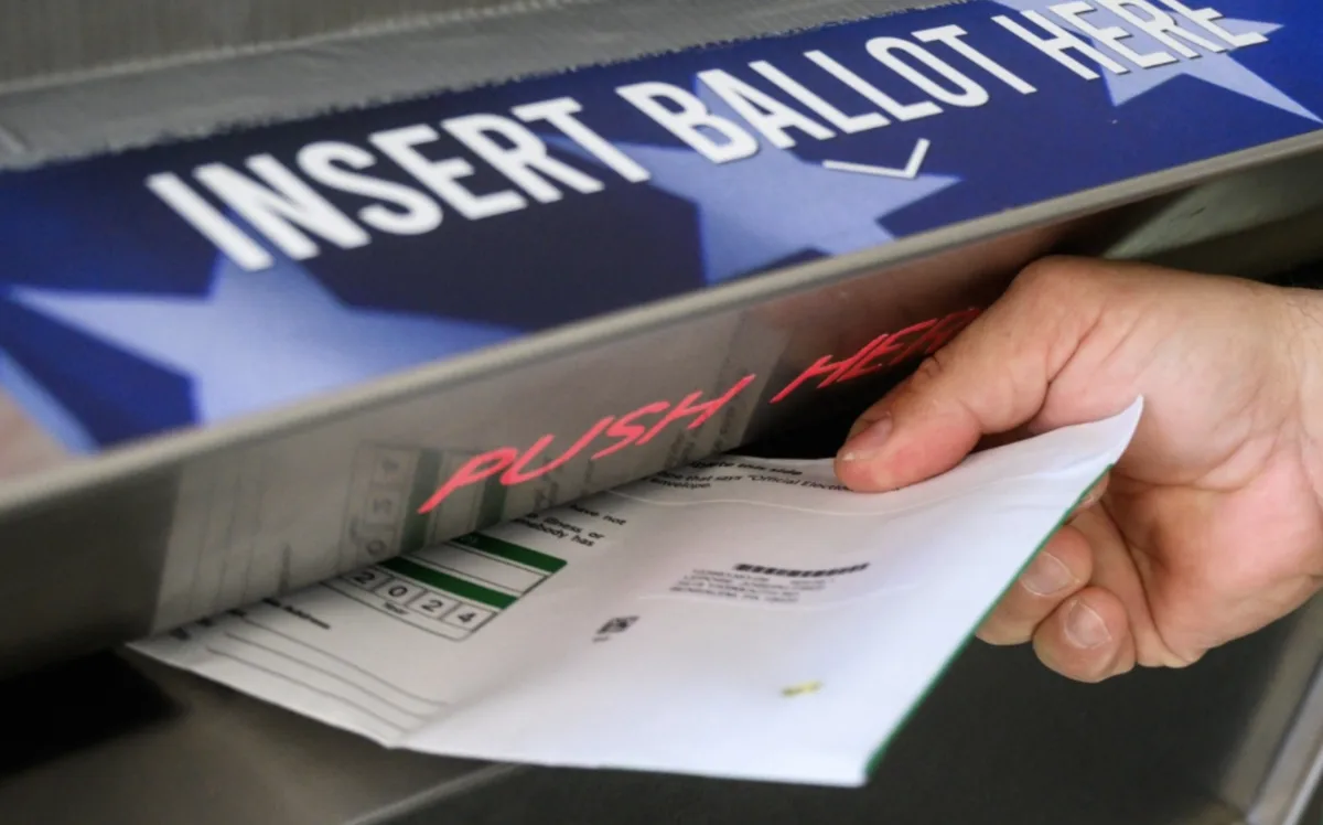 A close-up photo shows a person's hand inserting a 2024 election ballot into a ballot box. The box has a blue label with…