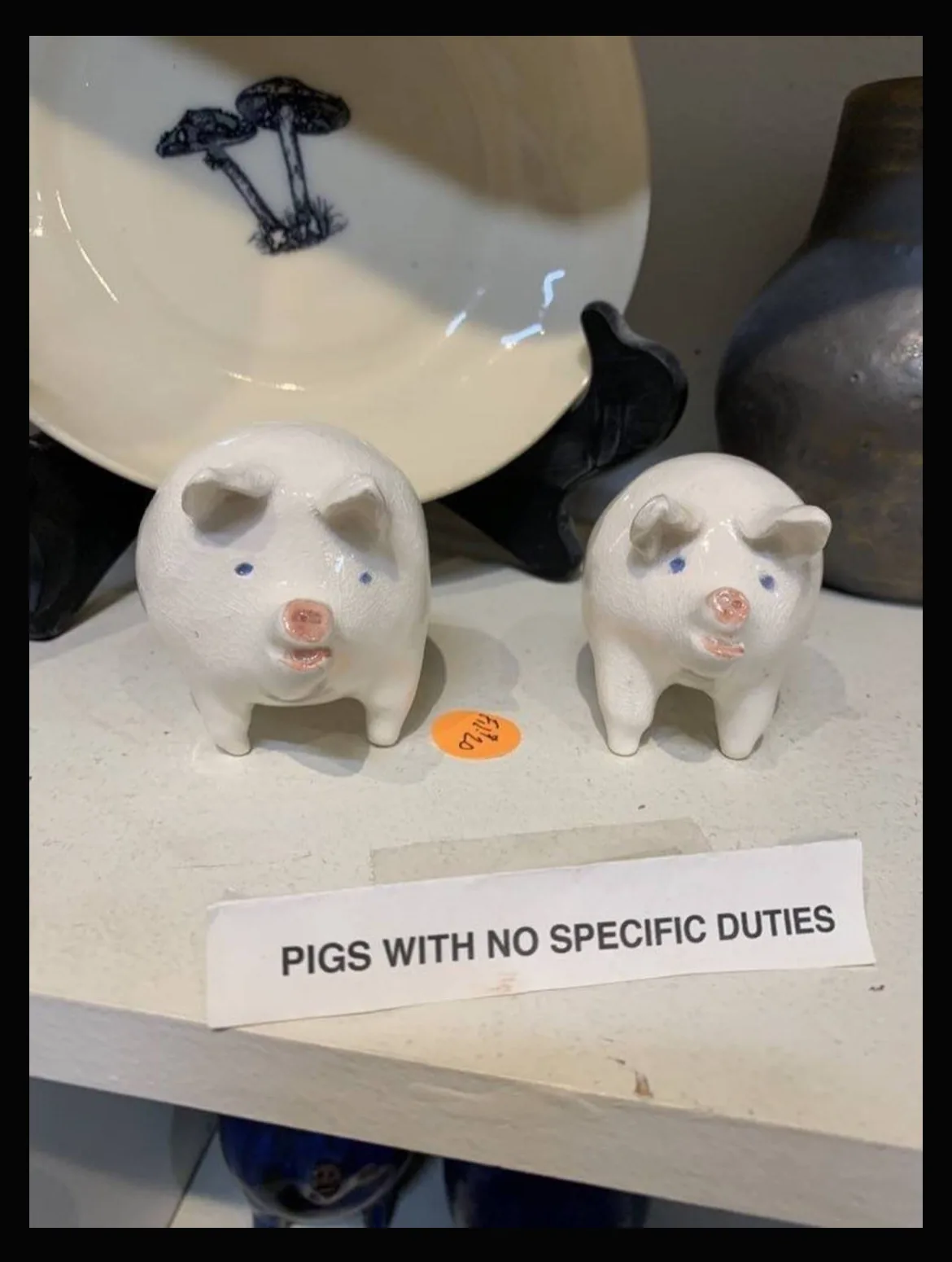 Two small white ceramic pig figurines with blue eyes and pink snouts stand side by side on a shelf, with a white paper label…
