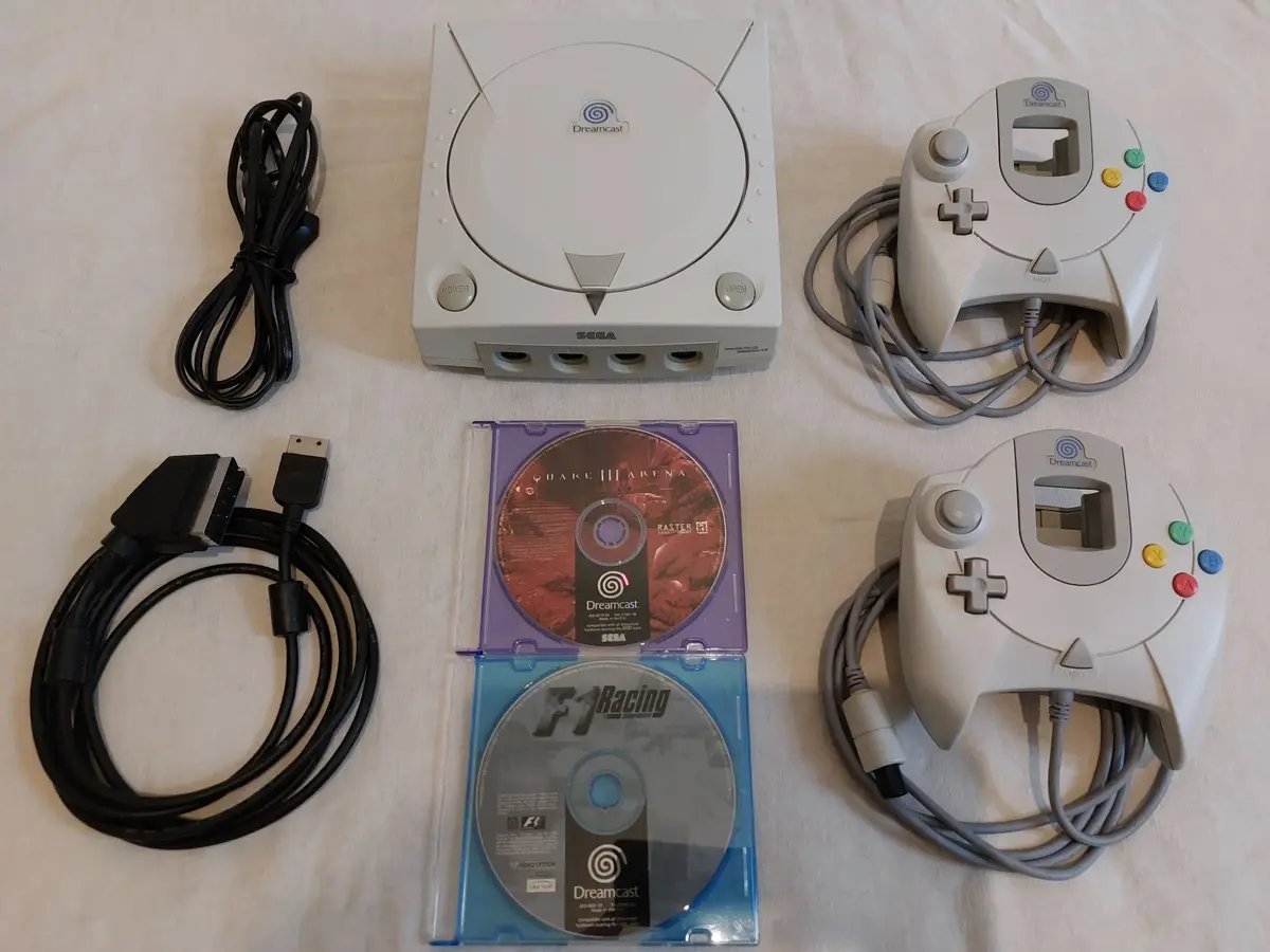 A top-down photo displays a white Sega Dreamcast console, two white Dreamcast controllers with gray cables, a black power…
