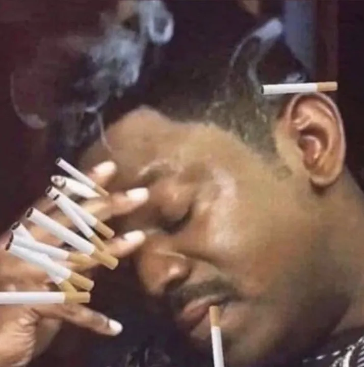 a man with his eyes closed and a weary expression has multiple cigarettes stuck in his hair, ear, and fingers, with smoke…