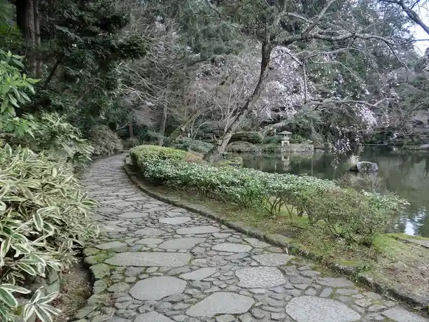 A serene stone path winds through a lush Japanese garden with cherry blossoms, green shrubs, and a pond with a stone…