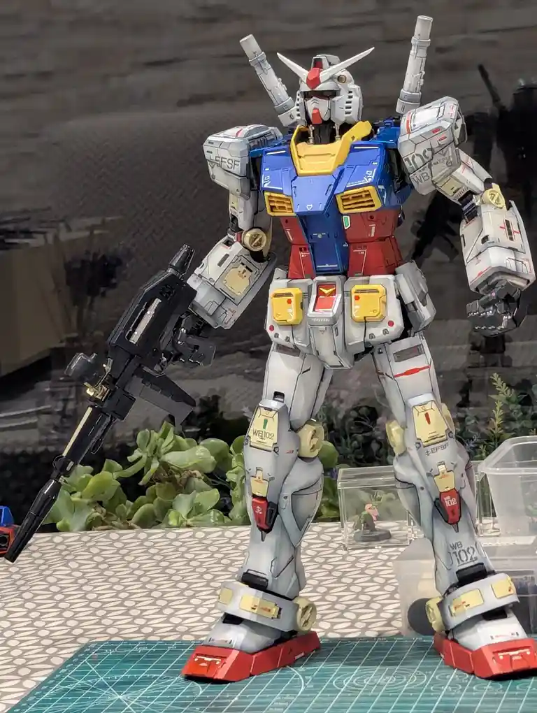 A detailed, custom-painted blue, red, and white Gundam model stands on a green cutting mat, holding a black rifle. The text…