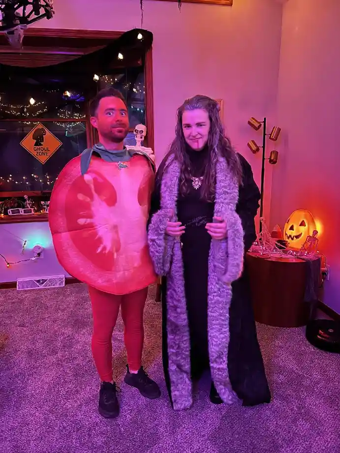 Two people in Halloween costumes stand indoors under purple lighting. The person on the left wears a large, red tomato…