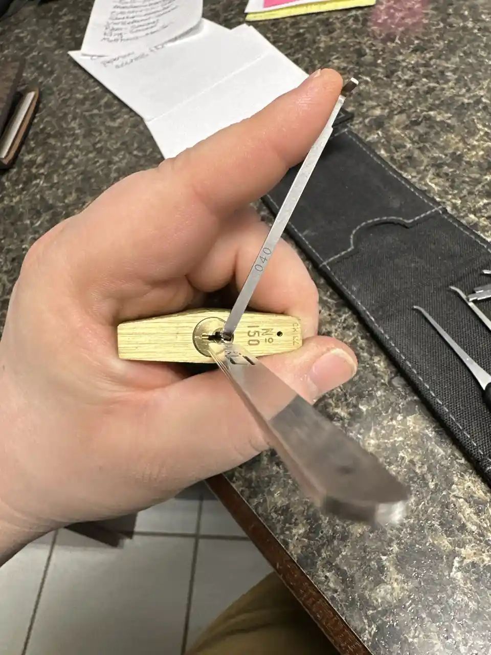A hand holds a brass padlock with a metal lockpick inserted into its keyway, while the pick's handle is blurred by motion…