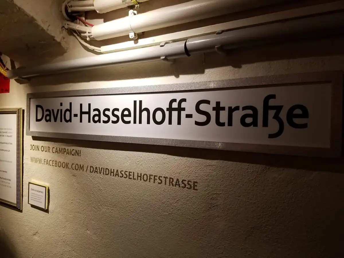 A street sign reading 'David-Hasselhoff-Straße' is mounted on a textured wall, with a campaign notice below it urging people…