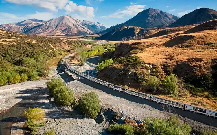 a scenic photograph shows a long passenger train traveling along a narrow track that crosses a wide, shallow riverbed in a…