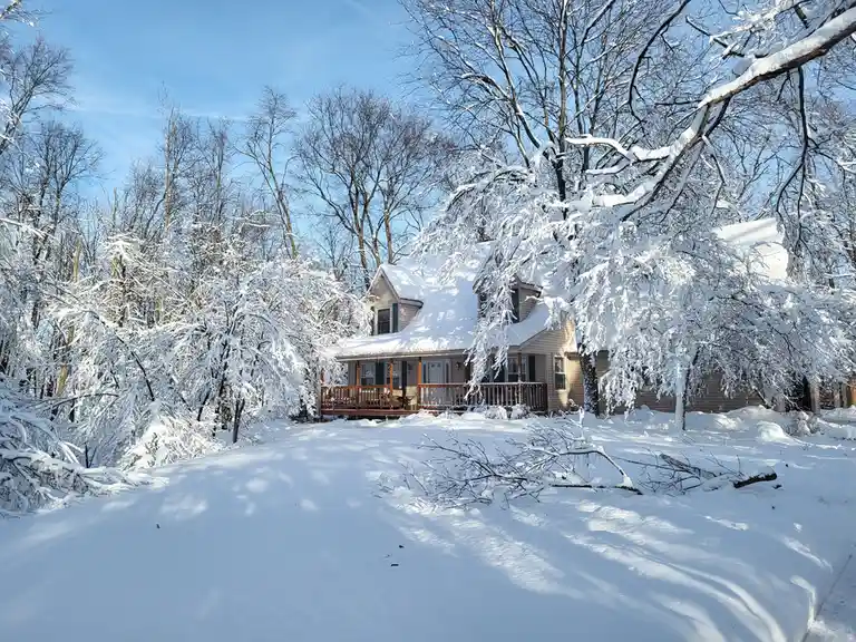 A house is surrounded by snow-covered trees under a clear blue sky, with snow blanketing the ground and roof. The overlaid…