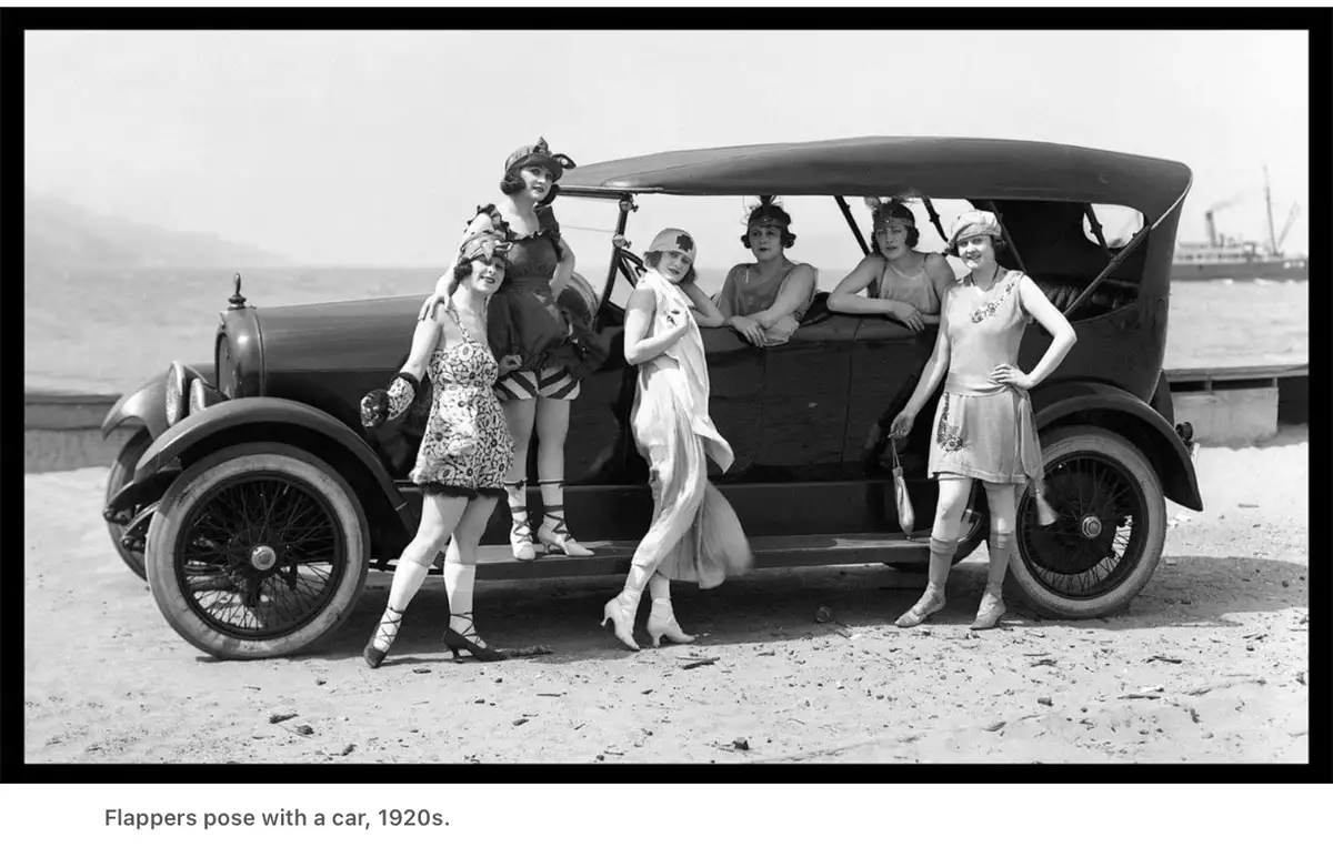 A vintage black-and-white photograph from the 1920s shows six young women, known as flappers, posing confidently around an…