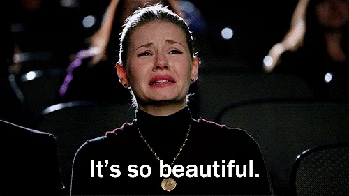 The image shows a woman in a dark theater wearing a black turtleneck and gold necklace, visibly crying with a distressed…