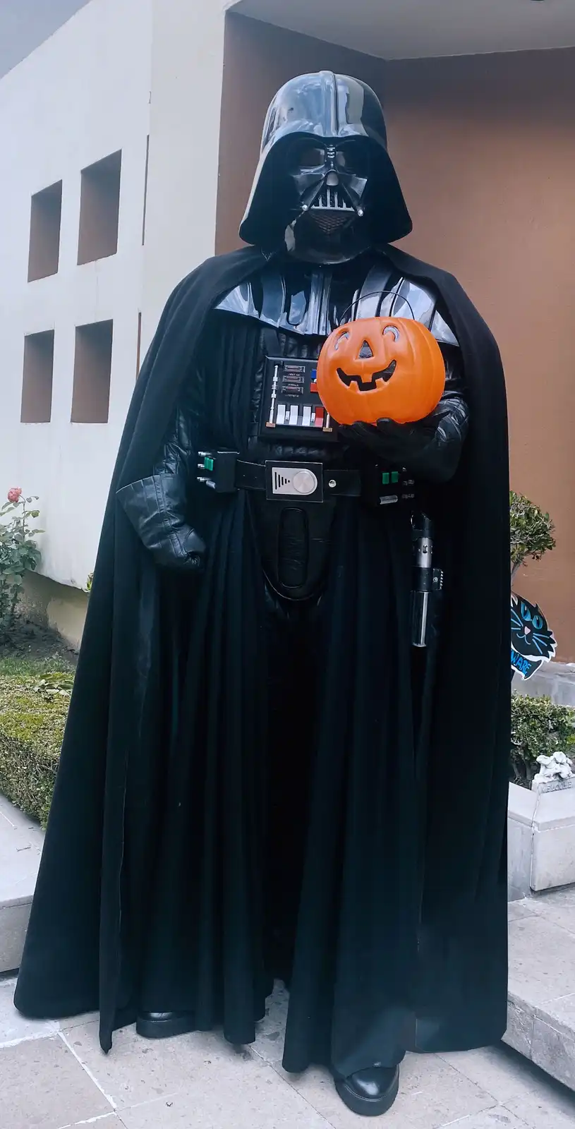 A person dressed as Darth Vader from Star Wars stands outdoors holding an orange plastic jack-o-lantern with a carved…