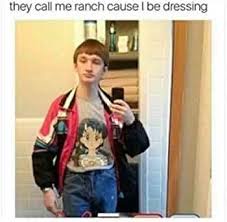 A young man takes a mirror selfie in a bathroom, wearing a graphic t-shirt, blue jeans, and a red-and-black jacket with…