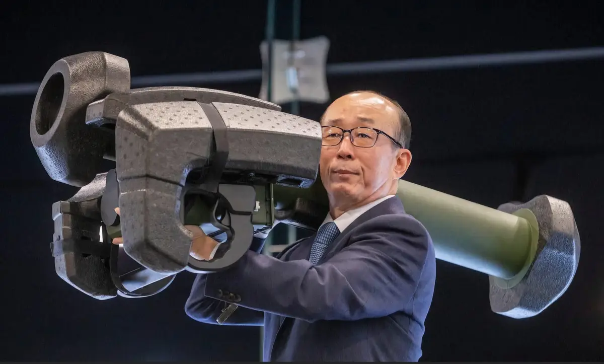 a man in a suit and glasses holds a large, heavy-looking cannon-like object, looking serious and determined. The overlaid…