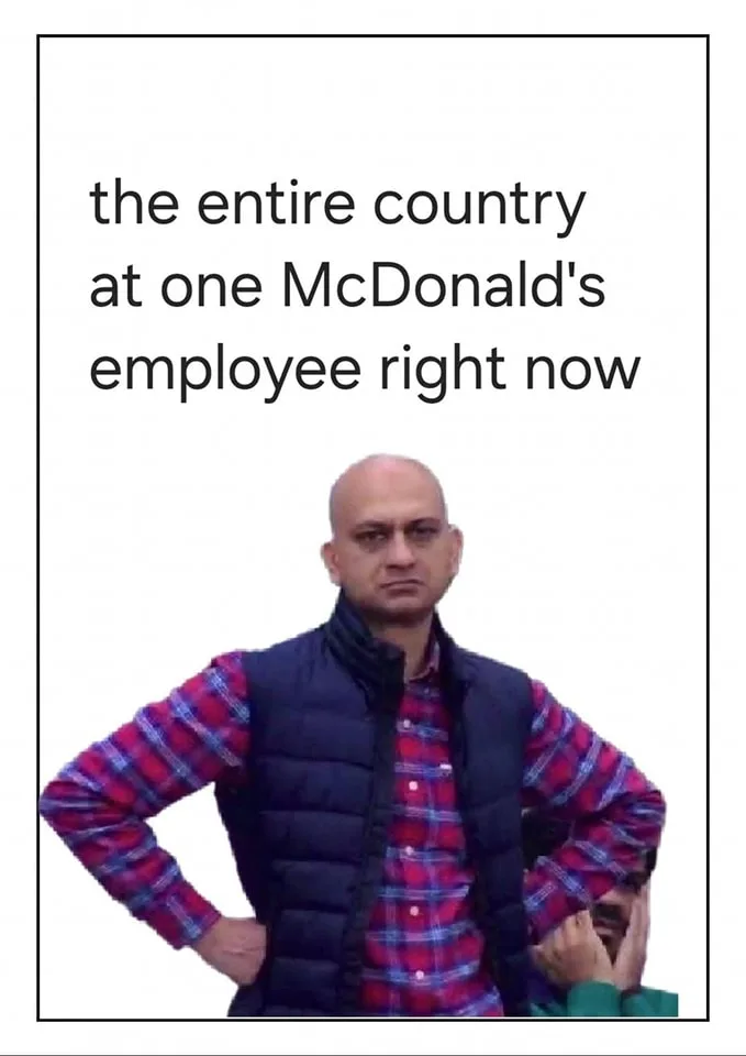 A bald man in a red plaid shirt and black vest stands with hands on hips, looking sternly at the camera. Behind him, another…