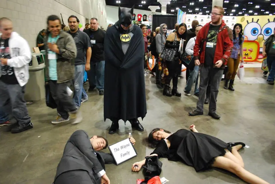 A person dressed as Batman stands over two people lying motionless on the floor at a convention, one holding a sign that…