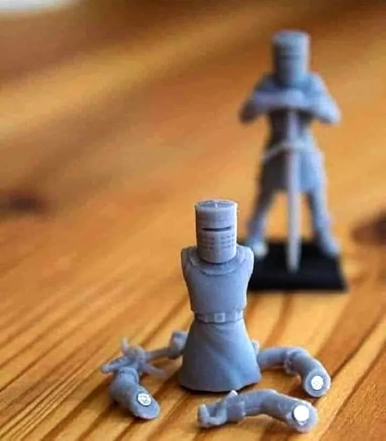 A close-up photo shows a gray 3D-printed knight miniature figurine on a wooden surface, with its arms detached and lying…