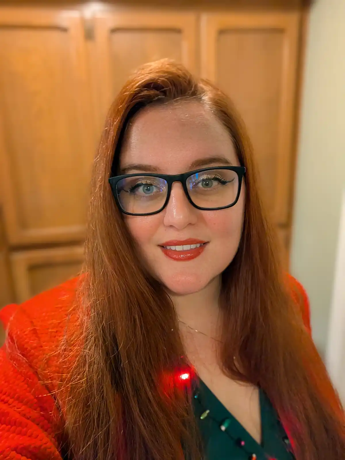 A woman with long red hair and black-rimmed glasses smiles at the camera while wearing a bright orange textured sweater over…