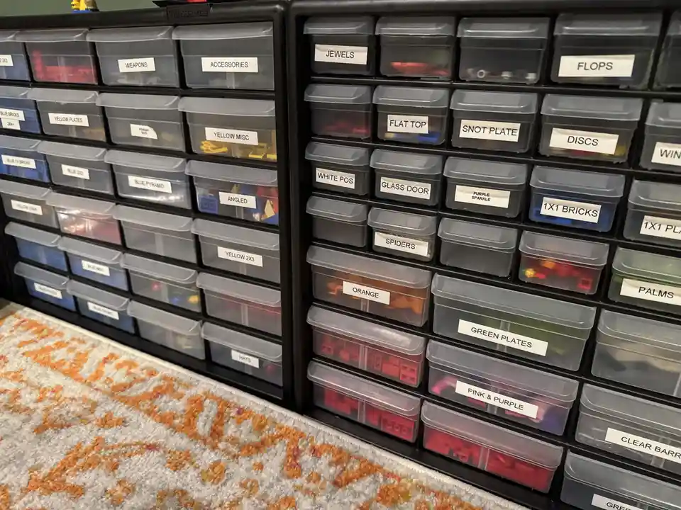 A wall of black plastic storage drawers filled with LEGO pieces, each drawer labeled with specific part names like 'JEWELS'…