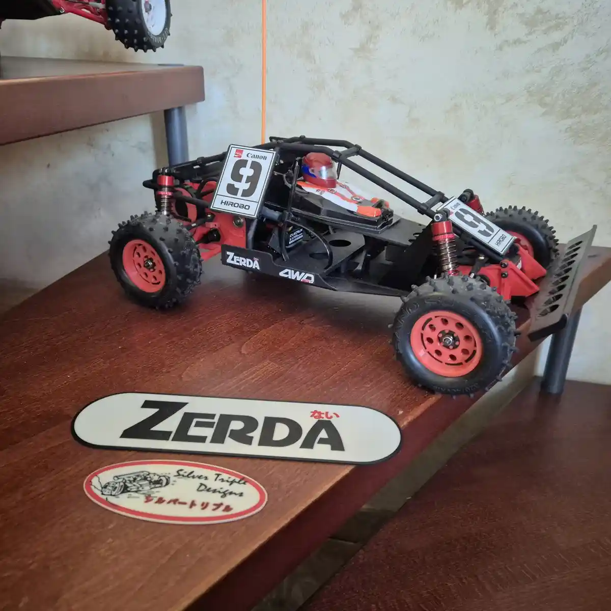 A red and black radio-controlled buggy with a driver figure sits on a wooden shelf, next to a ZERDA brand mat and a Silver…