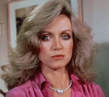 a still of a woman with voluminous blonde hair, wearing a pink blouse and pearl necklace, looking directly at the camera…