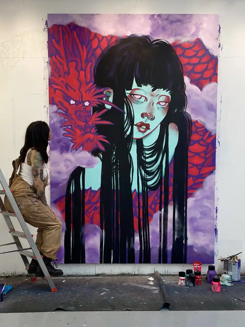 A person with tattoos sits on a ladder in an art studio, looking at a large, vividly painted mural. The mural depicts a…