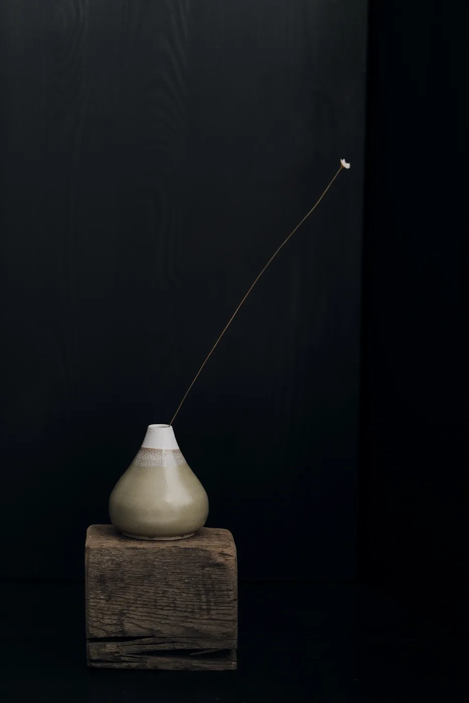A minimalist still life photograph shows a small, beige ceramic vase with a white rim, resting on a rough wooden block…