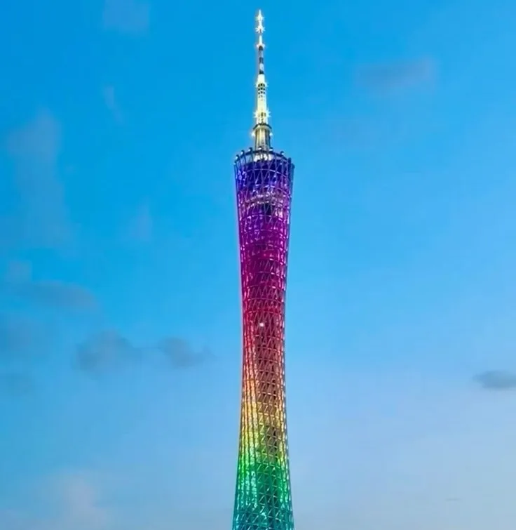A tall, slender tower with a lattice structure is illuminated with a full rainbow gradient of colors from top to bottom…