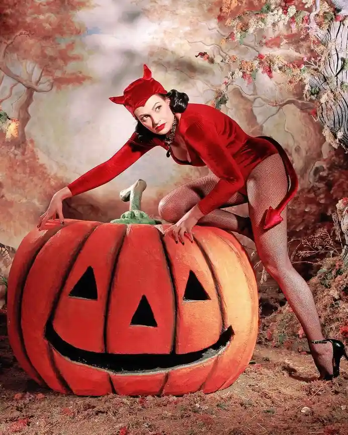 A woman in a red devil costume with horns and a tail poses seductively atop a large carved jack-o'-lantern. The background…