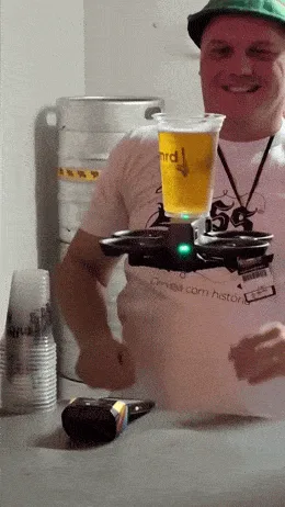 A man in a green hat and white t-shirt smiles while holding a drone carrying a beer cup at a bar. The drone flies away, then…
