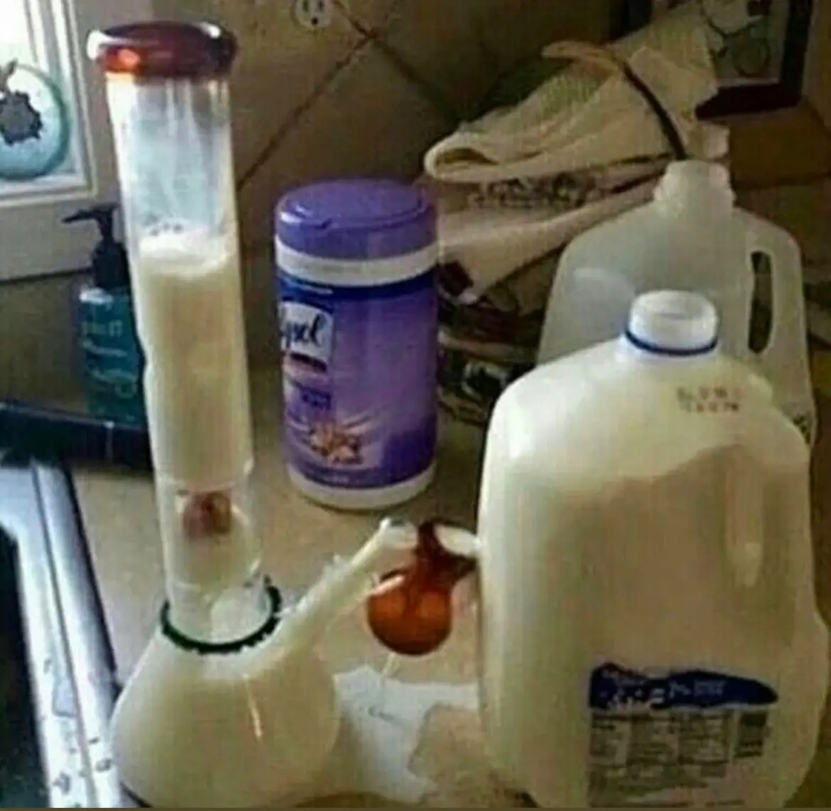 A bong is filled with milk and surrounded by milk jugs and baby wipes on a kitchen counter, creating a surreal and humorous…