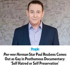A headshot of Paul Reubens, smiling, wearing a dark suit and light blue shirt, against a textured gray background. Below the…