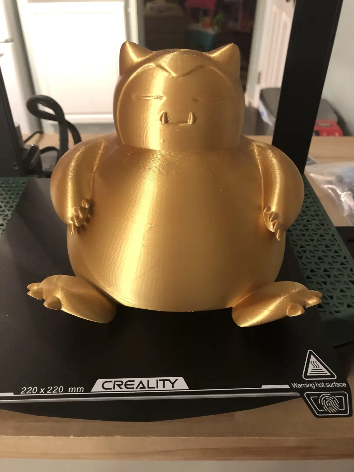 A golden 3D-printed statue of the Pokémon Snorlax sits on the build plate of a Creality 3D printer, which has visible…